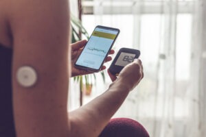 Woman comparing result of glucose level check on the traditional glucometer and on the mobile phone reading from modern technology remote sensor mounted on her forehand